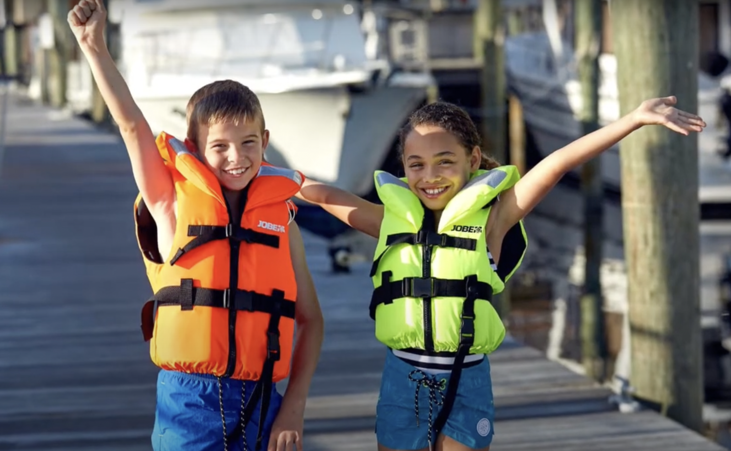 Two children wearing life jackets joyfully raise their arms while standing on a wooden dock. The boy on the left wears an orange life jacket, and the girl on the right wears a yellow one. Boats are visible in the background.